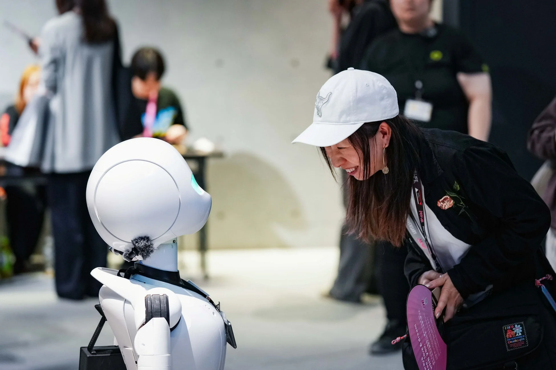 白いキャップをかぶった女性が、白いヒューマノイドロボットと笑顔で交流している様子を捉えた画像です。イベントか展示会のような場所で、人間とAIのポジティブな関わりを示しています。
