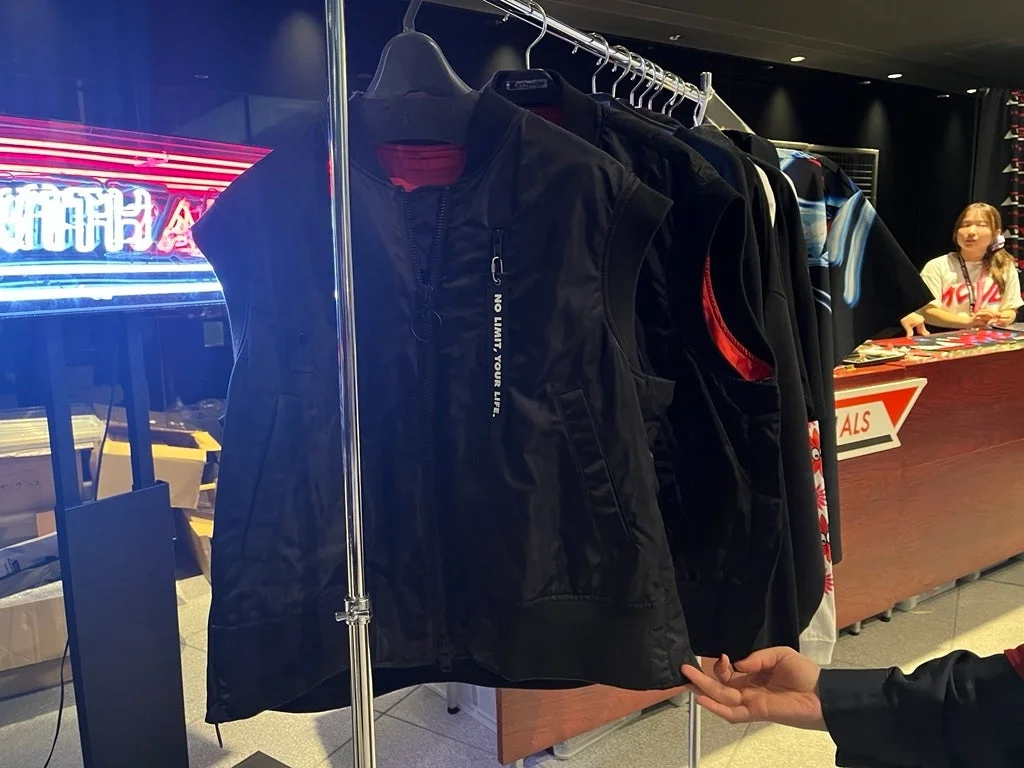 アパレルショップの店内で、ハンガーラックに吊るされた黒いベストやその他の衣料品が写っています。奥にはネオンサインがあり、店員らしき女性がカウンターに立っています。
