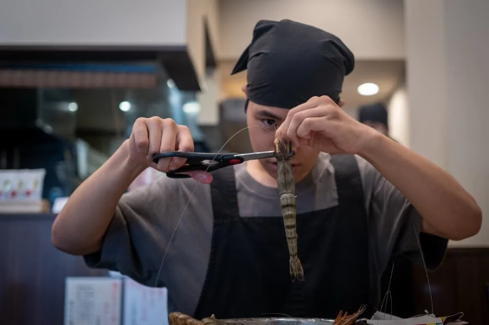 黒いバンダナとエプロンを着用した人物が、大きなエビの背わたをハサミで丁寧に取り除いている様子。厨房のような場所で料理の下準備をしている場面です。