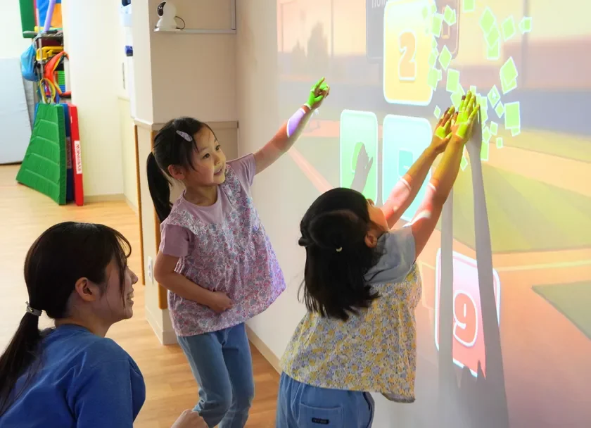 2人の女の子が壁に投影されたインタラクティブなゲームで遊んでいる様子です。数字が表示され、子供たちは手を伸ばして楽しんでいます。横で大人が見守っており、明るい室内で教育的な遊びが行われているようです。