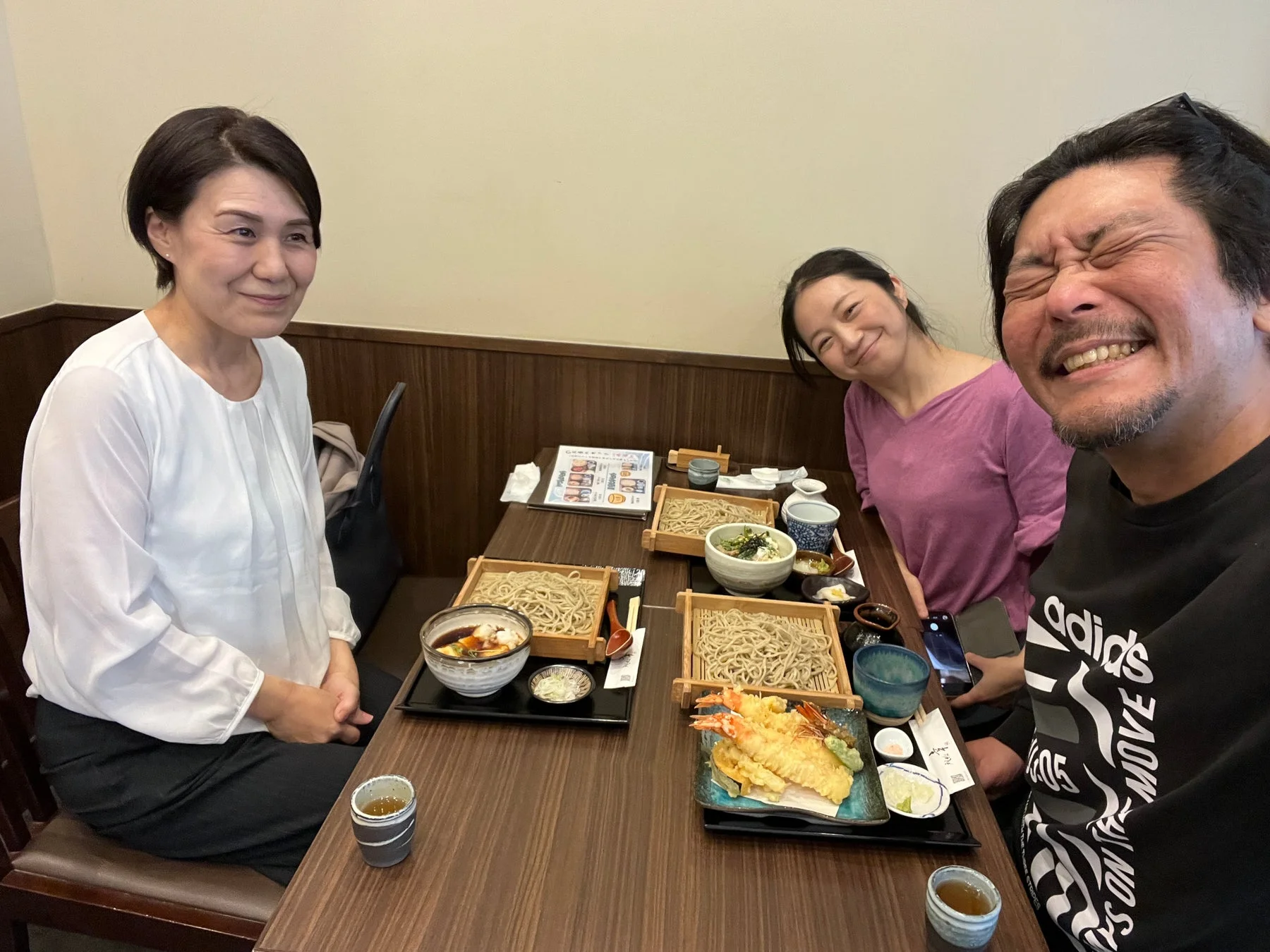 3人の人々がレストランで楽しそうに食事をしている様子です。テーブルには蕎麦と天ぷらが並び、皆笑顔で和やかな雰囲気です。