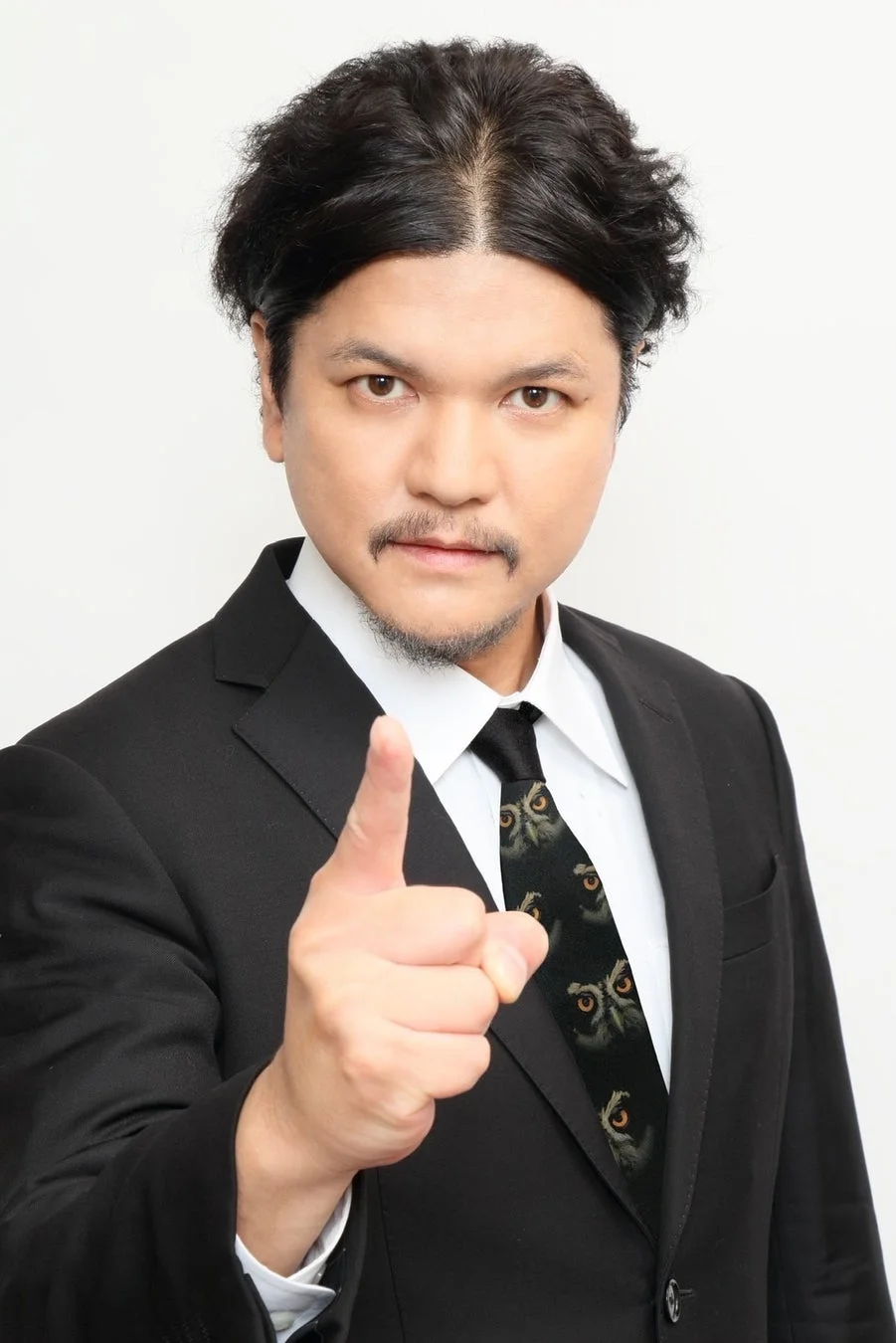 黒いスーツとフクロウの目の柄のネクタイを着用した男性が、正面に向かって人差し指を指しているポートレート。