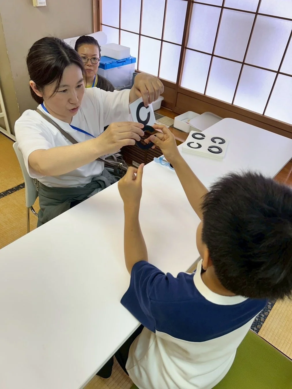 女性が子供に対してランドルト環を使った視力検査のような活動を行っています。テーブルには複数のランドルト環が置かれており、後ろには別の大人が見守っています。和室のような空間で行われている様子です。
