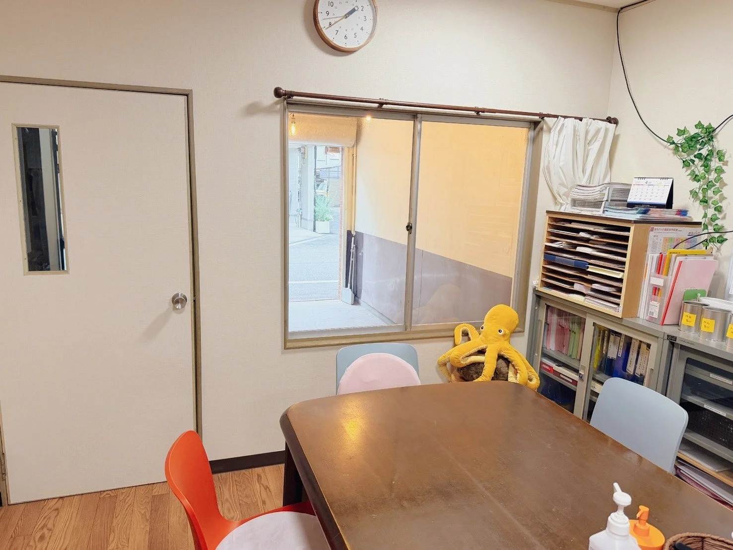 室内の一室を写した画像です。大きな木製テーブルと椅子が複数置かれ、窓からは外の景色が見えます。右側には書類棚があり、黄色いタコのぬいぐるみも置かれています。壁には時計がかかっており、機能的ながらも温かみのある空間です。
