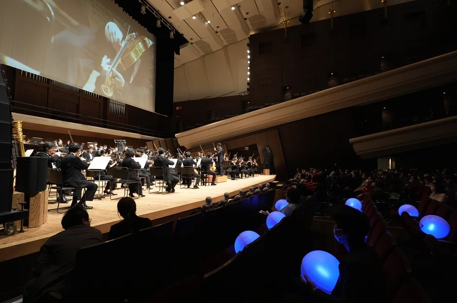 コンサートホールでオーケストラが演奏している様子。舞台奥のスクリーンにはバイオリンの演奏が映し出され、客席では観客が青く光る風船を持っている。