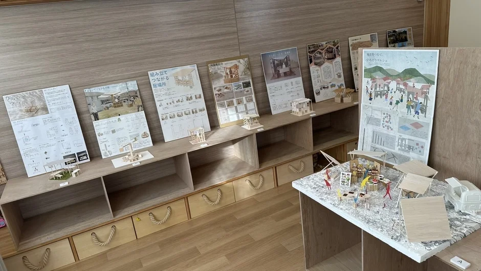 木製の建築模型と解説パネルが並べられた展示会の様子。組み立て式の居場所やいろどりマルシェといったテーマで、仮設的なコミュニティ空間や商業スペースのデザイン案が展示されており、学生の設計作品展のようである。