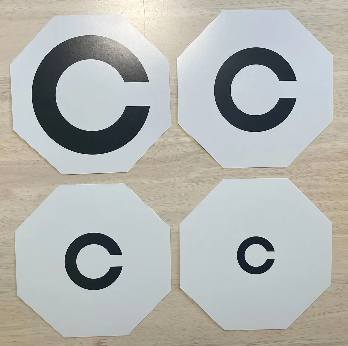 木目調の背景に、白地に黒いCの文字が書かれた八角形のカードが4枚並べられています。それぞれのCの文字は大きさが異なり、視力検査などで使用されるランドルト環に似ています。