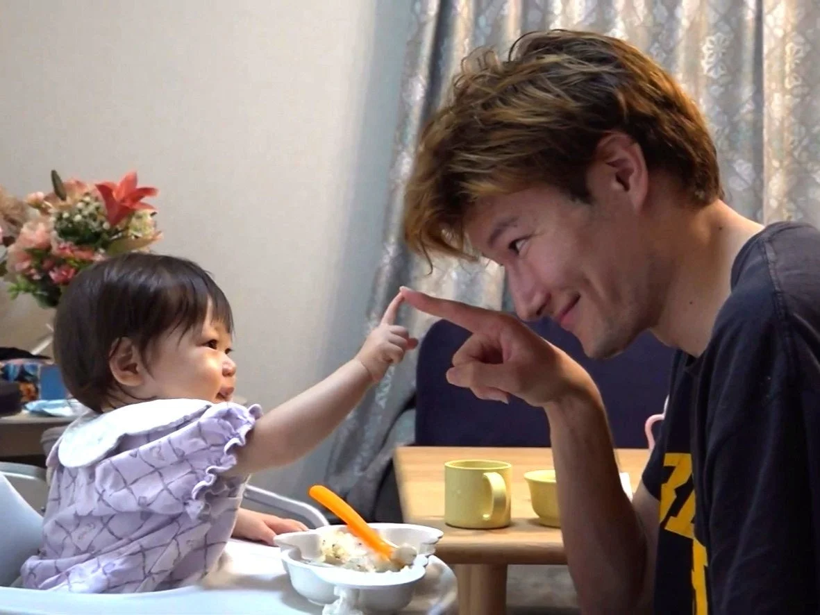 ハイチェアに座った赤ちゃんが、笑顔の男性の指にそっと触れている心温まる親子の触れ合いの瞬間です。食事中の日常の一コマが写し出されています。