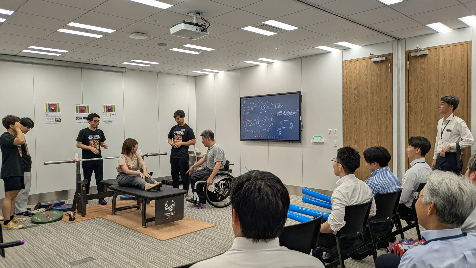 オフィスでパラ・パワーリフティング体験会に参加する社員たち。スクリーンには「怪力決定戦!」の文字