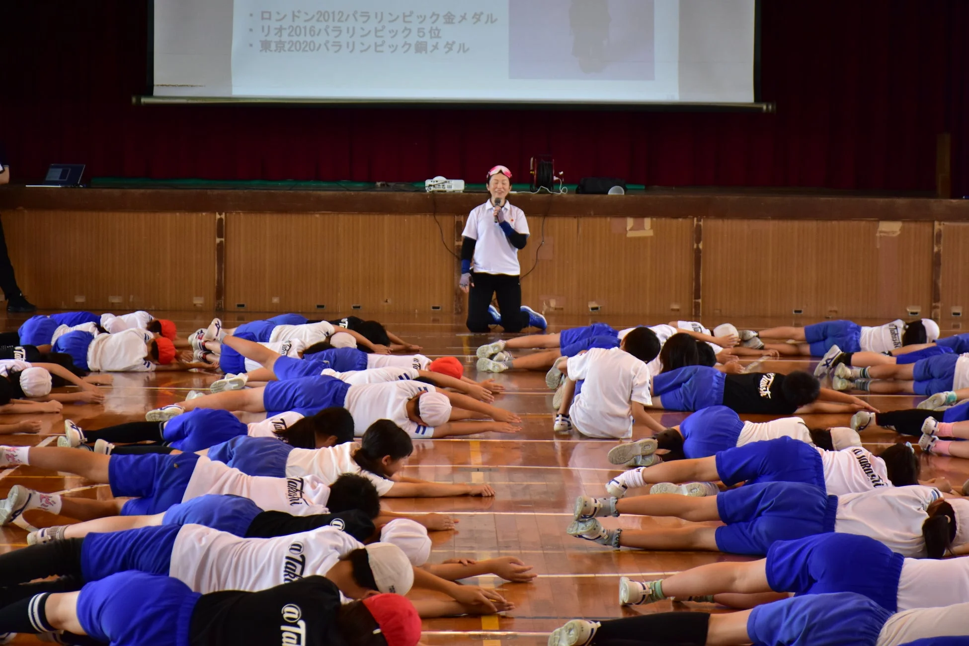 体育館で、多くの生徒が体操服姿で床に伏せている。前方にはマイクを持つ講師がおり、背後のスクリーンにはパラリンピックでのメダル獲得実績などが表示されている。運動指導や講演会が行われている様子。