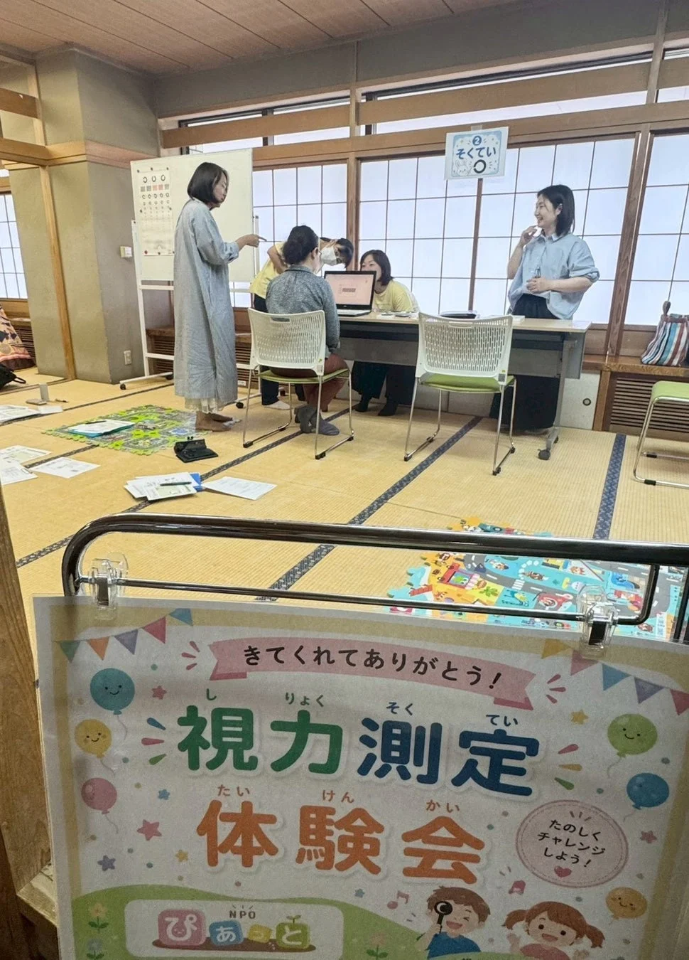 畳の部屋で開催されている「視力測定体験会」の様子です。参加者が視力検査を受けている横で、スタッフが説明を行っています。明るく親しみやすい雰囲気のイベントで、NPO「ぴあと」が主催しているようです。