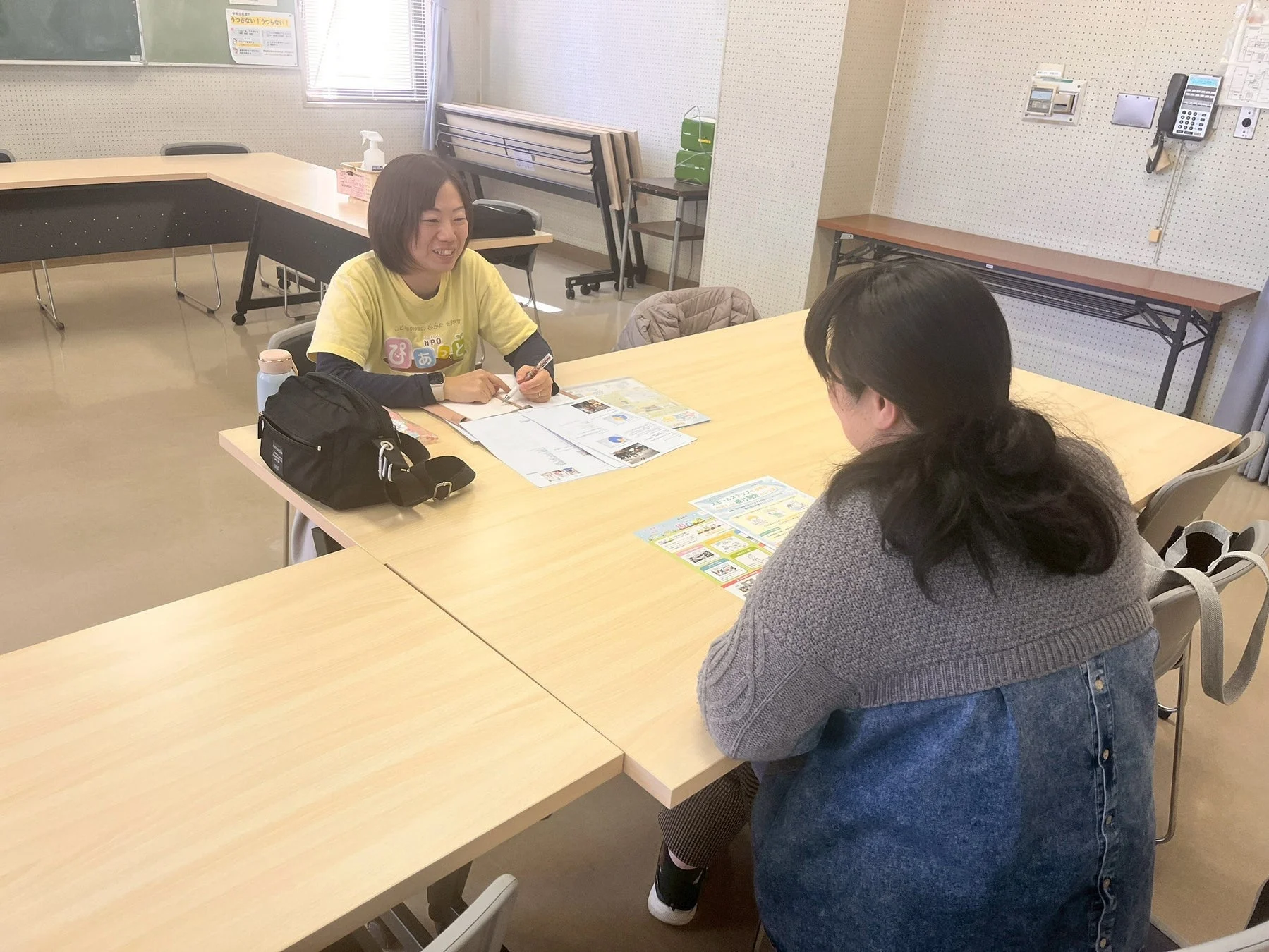会議室のような場所で、2人の女性がテーブルを囲んで対話しています。左の女性は笑顔で書類に記入しており、右の女性と相談している様子です。