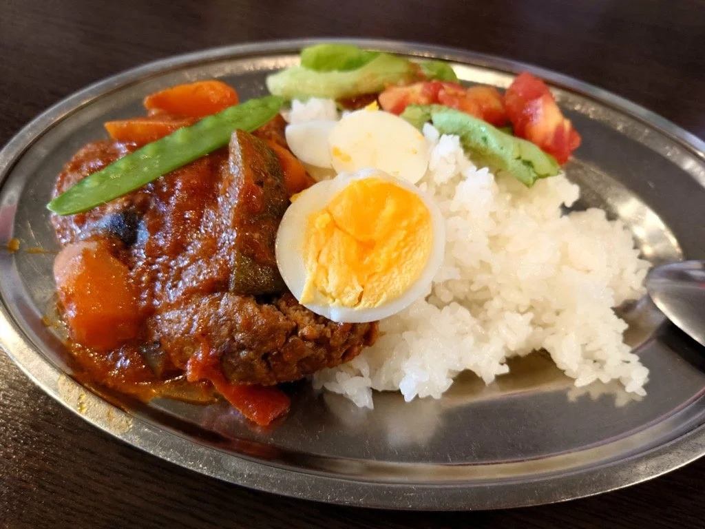 金属製の皿に盛られた洋食プレート。ライス、煮込みハンバーグのようなメイン料理、ゆで卵、レタスやトマトなどの野菜が彩り豊かに盛り付けられています。