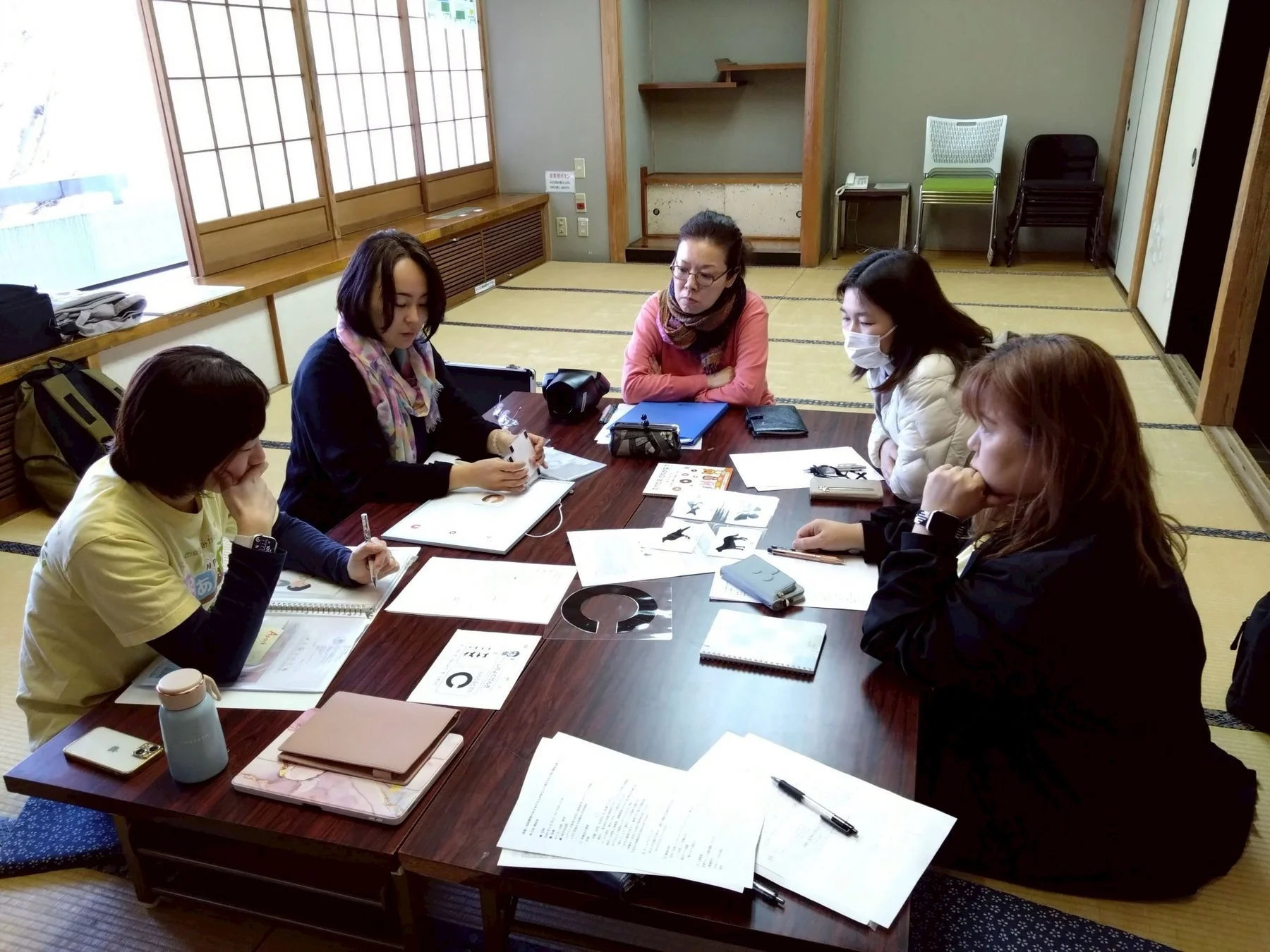 和室で5人の女性が低いテーブルを囲み、書類やノートを広げて真剣に話し合ったり作業したりしています。会議やワークショップのような雰囲気で、共同で何かに取り組んでいる様子が伺えます。