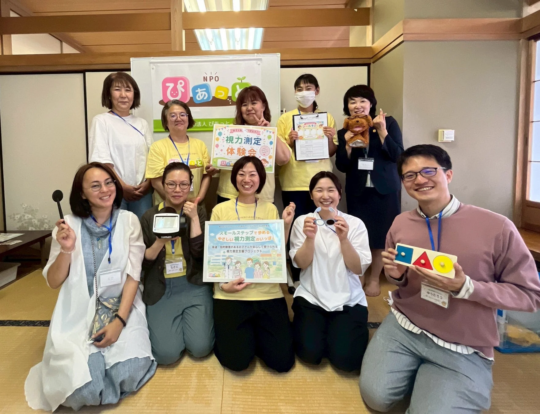 NPO「ぴあっと」が主催する、発達・知的障害のあるお子さん向けの「やさしい視力測定体験会」の集合写真です。参加者たちは笑顔で、視力測定に関する看板や器具、おもちゃなどを手にしています。畳敷きの和室で和やかな雰囲気の中、プロジェクトの成功を祝っているようです。