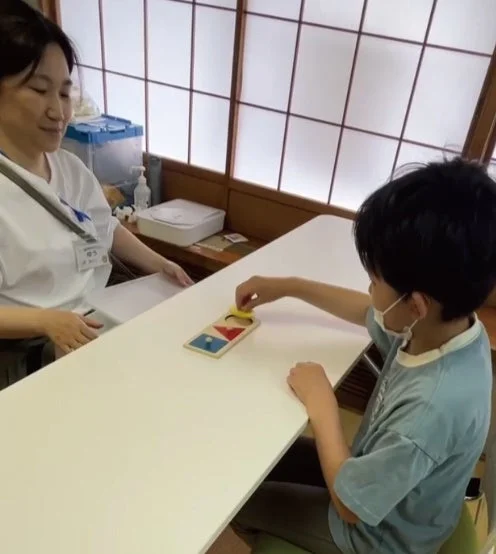 室内で、女性が子どもが木製パズルに取り組む様子を見守っています。子どもはマスクを着用し、集中してパズルをはめ込んでおり、学習や療育活動の場面と考えられます。
