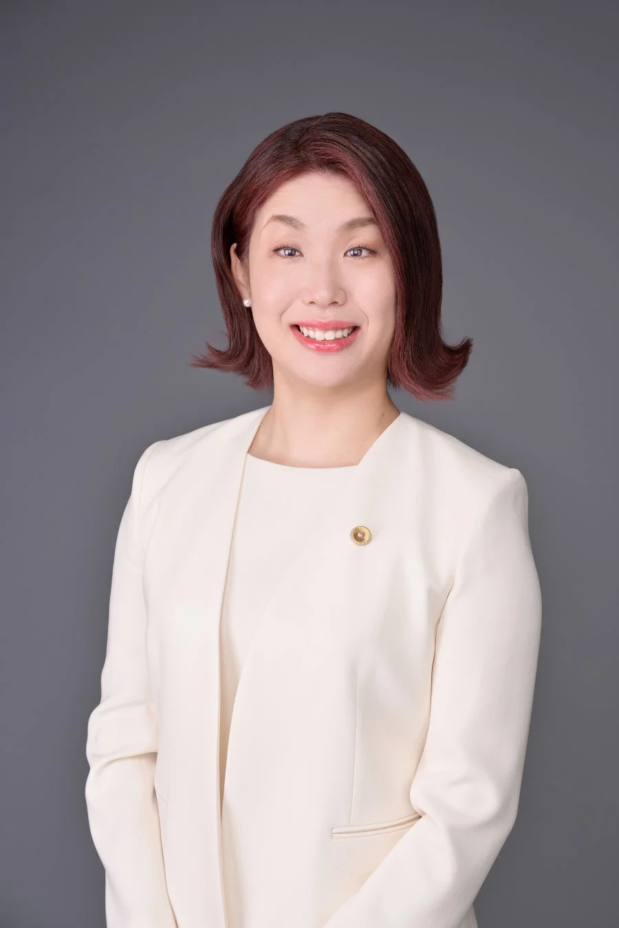 白いジャケットを着用し、笑顔でカメラを見つめるアジア人女性のポートレートです。ショートヘアで、左の襟元には金色のピンがつけられており、プロフェッショナルな印象を与えます。