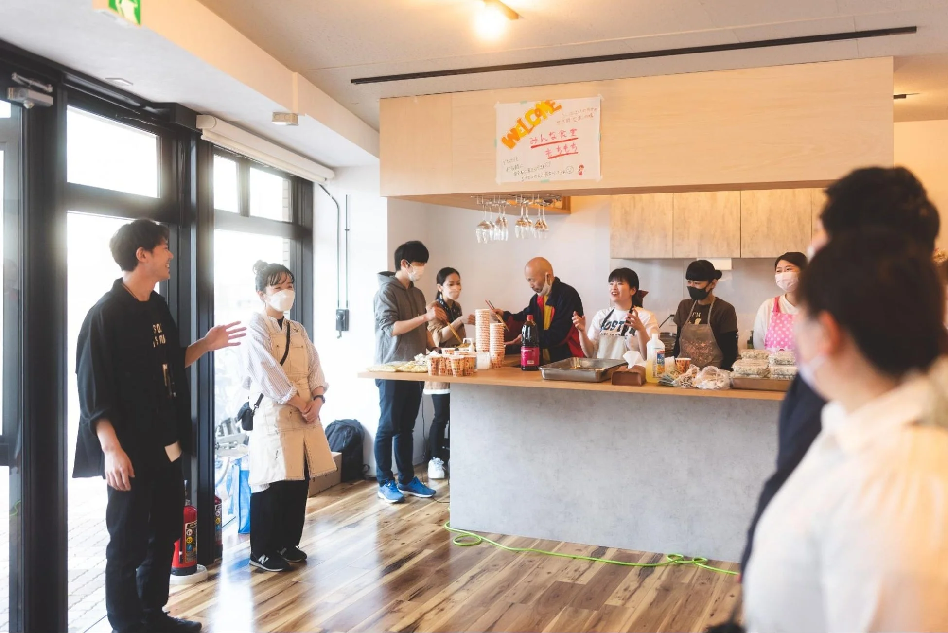 明るくモダンな空間で、老若男女がカウンターを囲み、食事の準備や提供をしている様子。壁には「みんな食堂」の歓迎サインがあり、地域の人々が集う温かい交流イベントが開催されている。