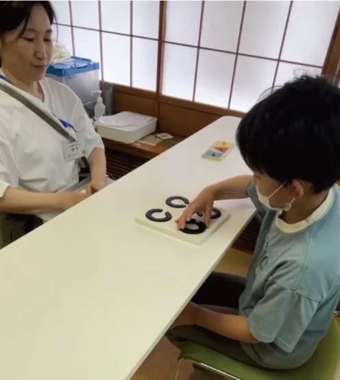 女性と子供が白いテーブルに向かい合っています。子供はマスクを着用し、黒いリングのパズルや知育玩具で遊んでいます。女性は指導者かセラピストのようで、子供の活動を見守っています。療育や学習の場面を捉えた一枚です。
