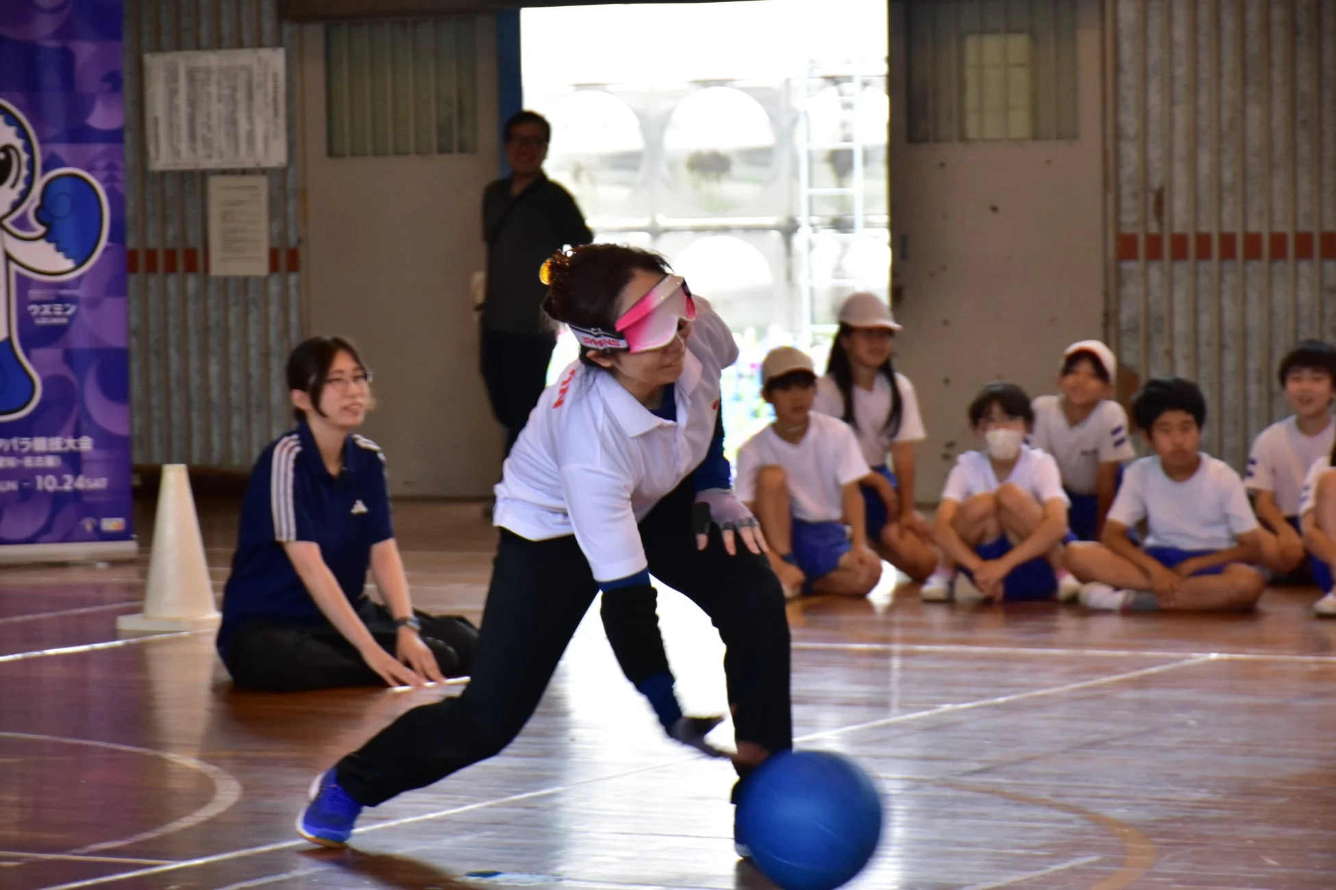 体育館でブラインドゴーグルをつけた人が青いボールを転がしているパラスポーツの様子。周りには複数の子供たちが座って見学しており、体験イベントか教室のようだ。