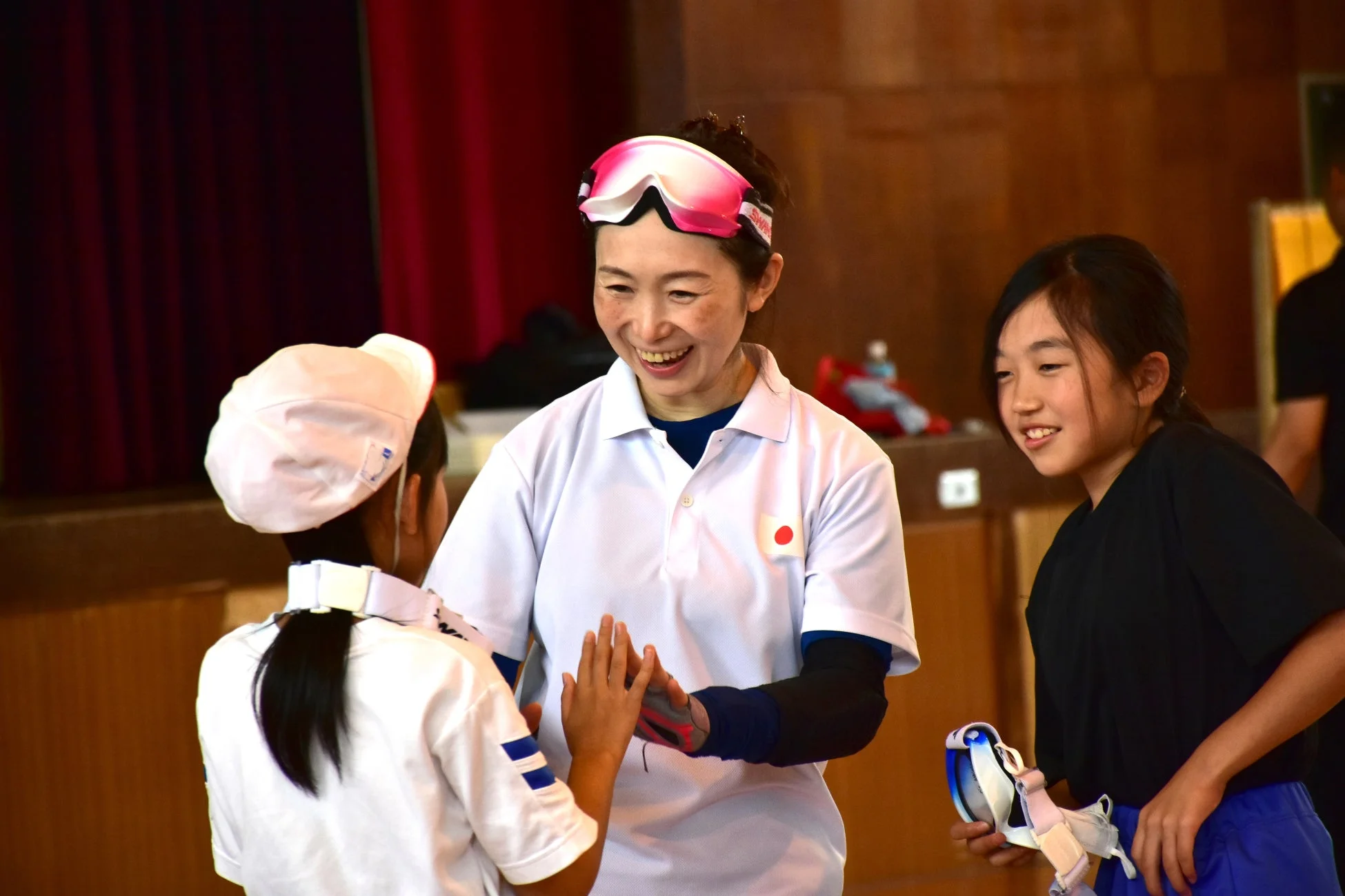 ゴーグルを頭につけた女性が、二人の子供たちと笑顔で交流している様子。体育館のような屋内で、スポーツ指導やレクリエーション活動が行われている場面です。