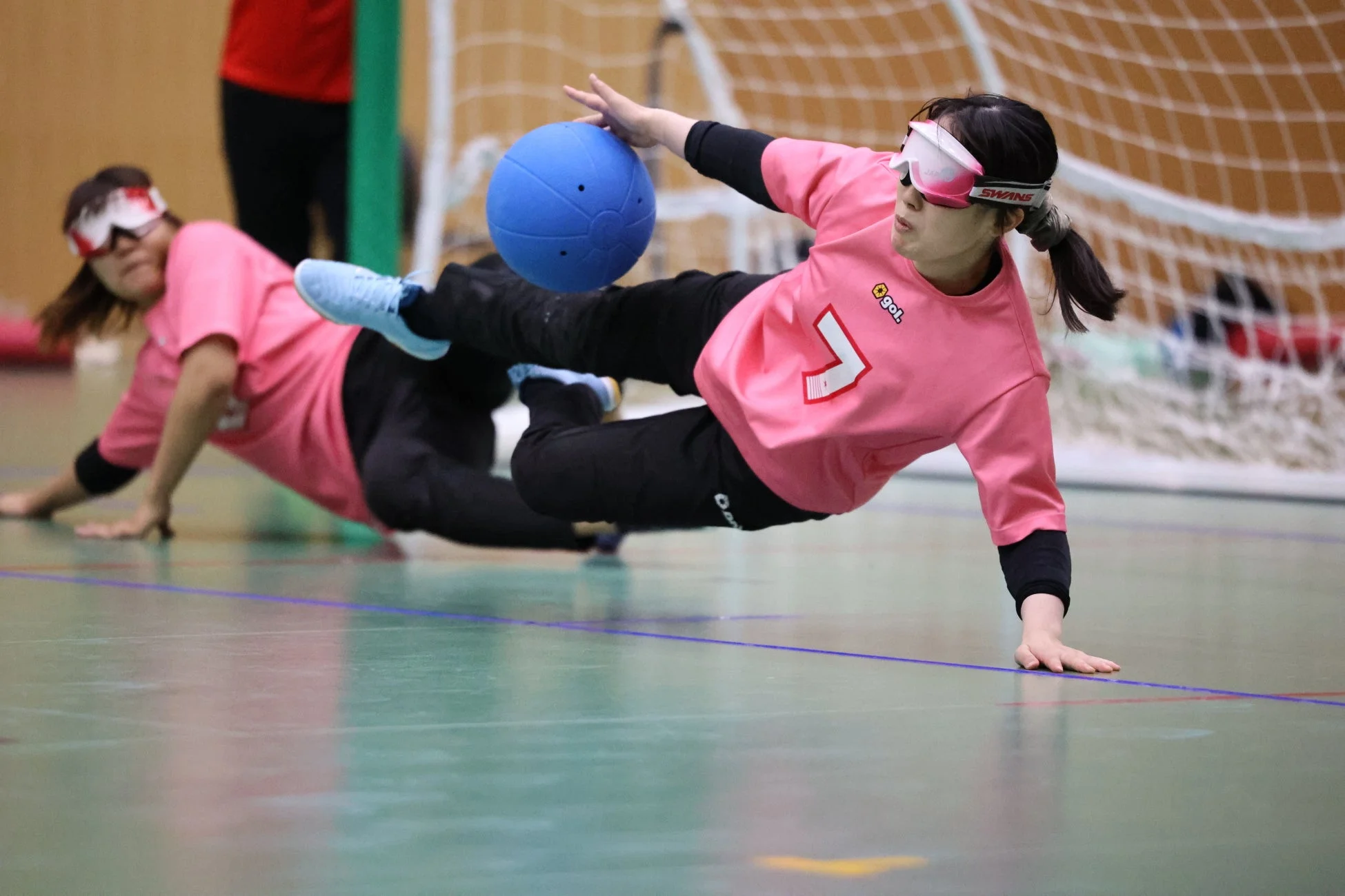 ピンクのユニフォームとアイシェードを着用した2人の女性ゴールボール選手が、青いボールをブロックしようとダイブしている瞬間を捉えた画像です。手前の選手は空中に浮き、真剣な表情でボールに集中しています。視覚障がい者スポーツの激しさと集中力が伝わる一枚です。