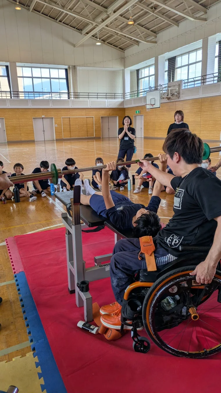 体育館で車椅子を使用する男性がベンチプレスをする子供を補助している様子