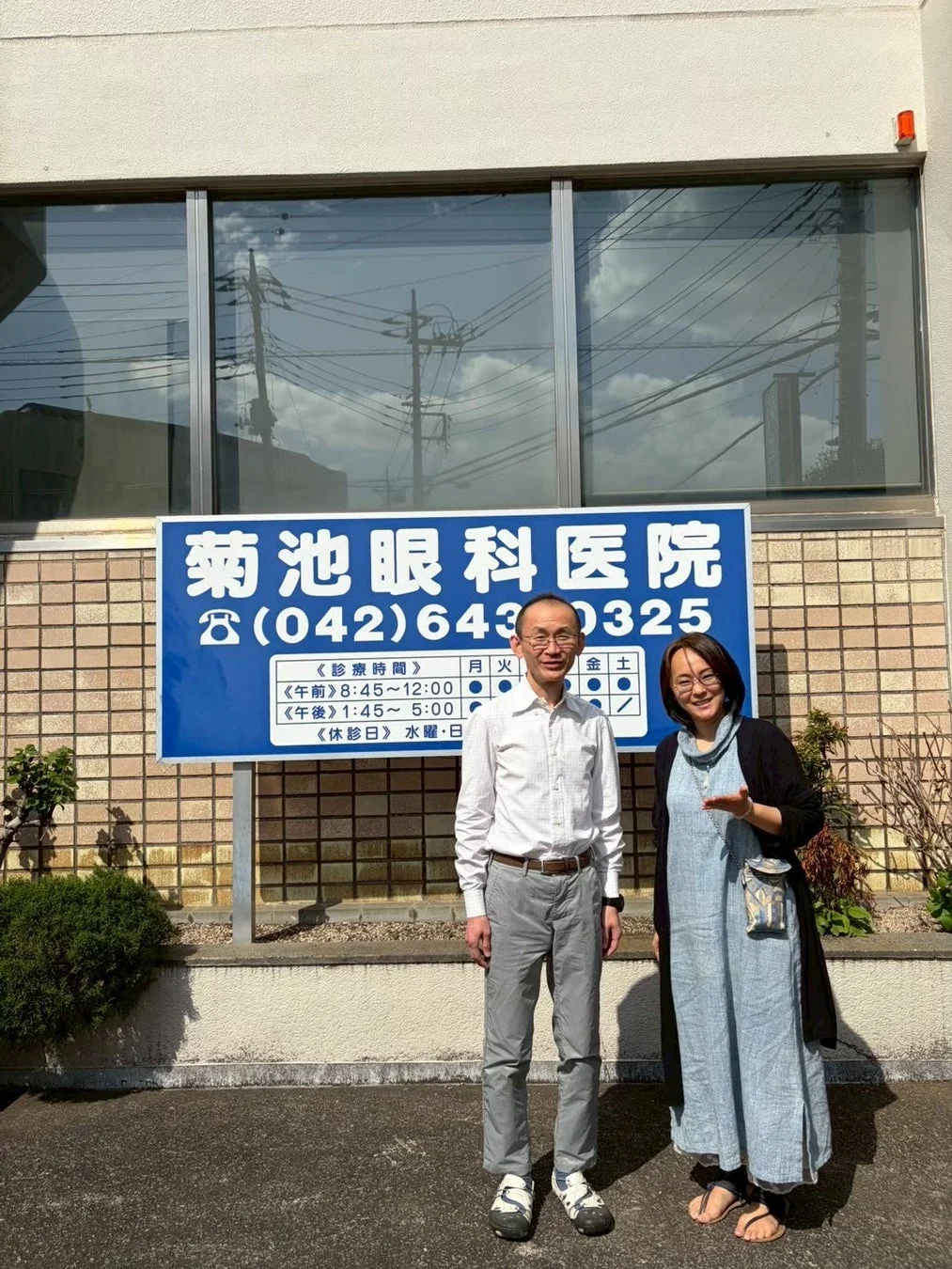 菊池眼科医院の看板の前で、男女2人が笑顔で立っている様子を捉えた写真です。看板には診療時間や電話番号が明記されており、クリニックの外観が伺えます。