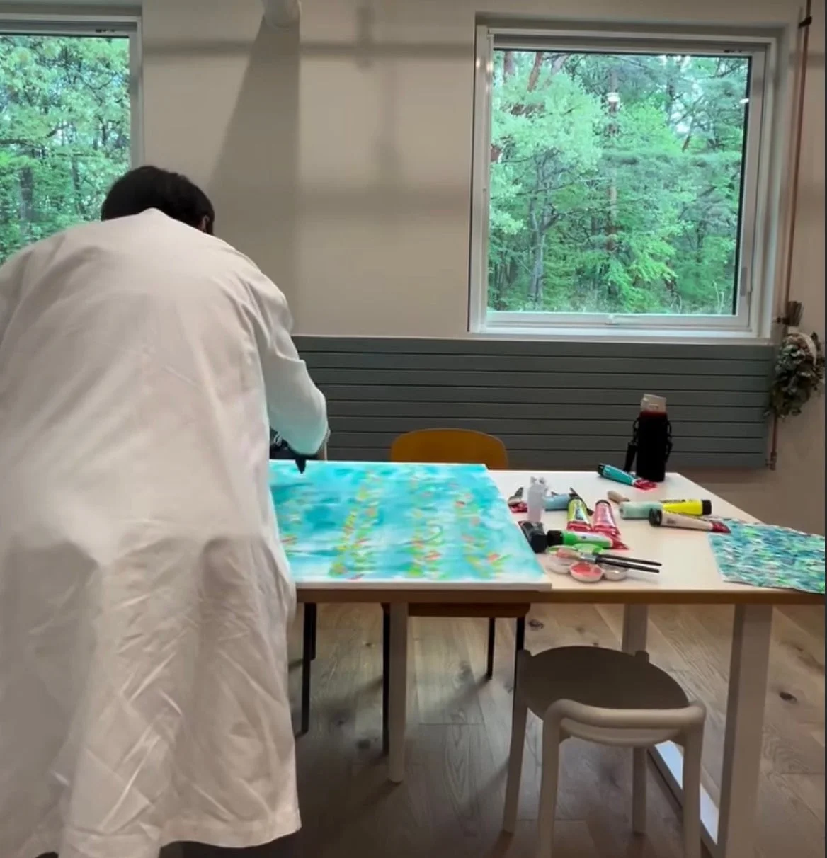 森の見える窓がある明るい部屋で、白い服を着た人物がキャンバスに抽象画を描いています。テーブルには絵の具や筆などの画材が並び、創作活動に集中している様子が伺えます。
