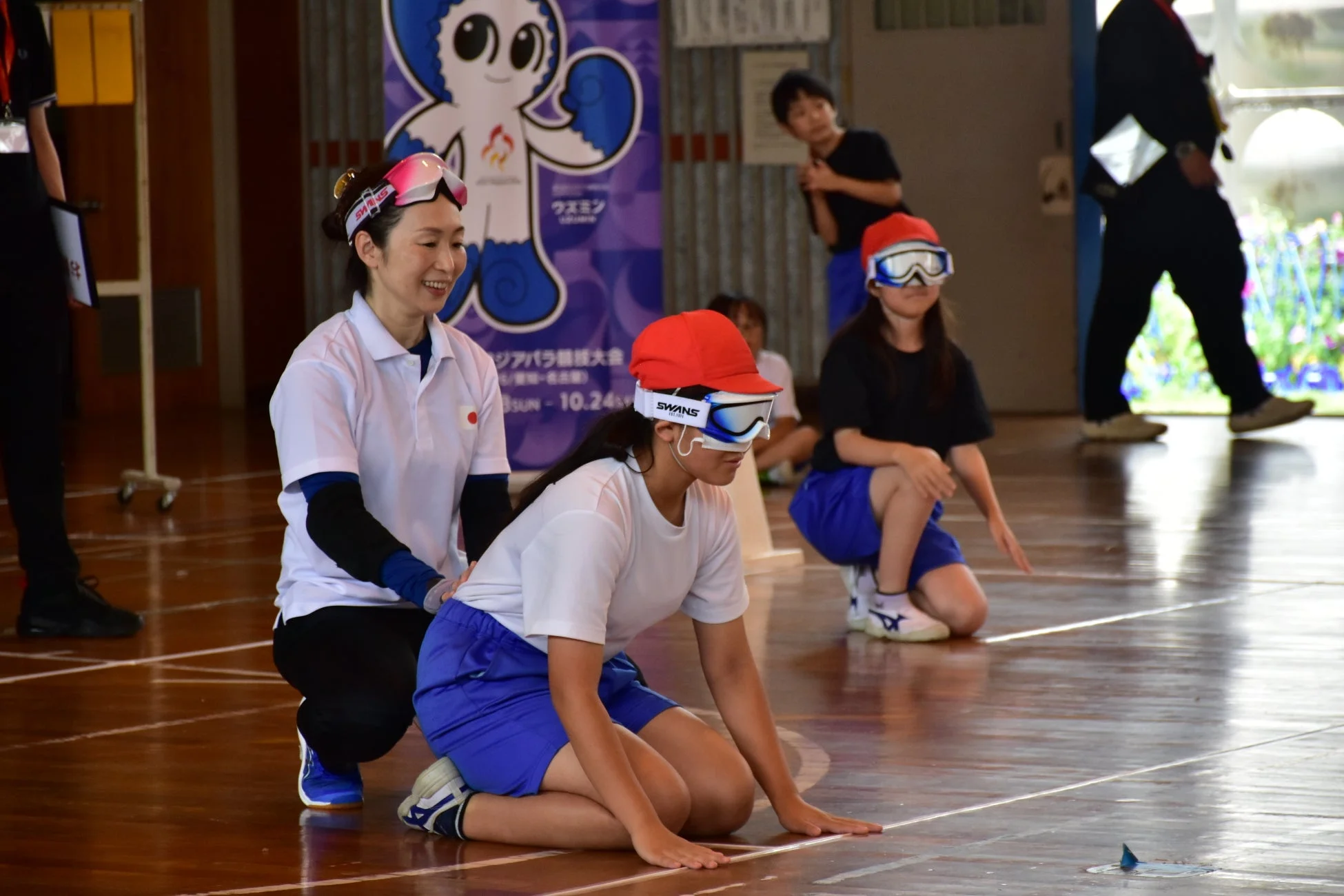 体育館で行われているパラスポーツ体験イベントの様子。目隠しゴーグルをつけた参加者を笑顔の女性がサポートしており、子供たちが集中して活動している。背景には「アジアパラ競技大会」のバナーが見える。