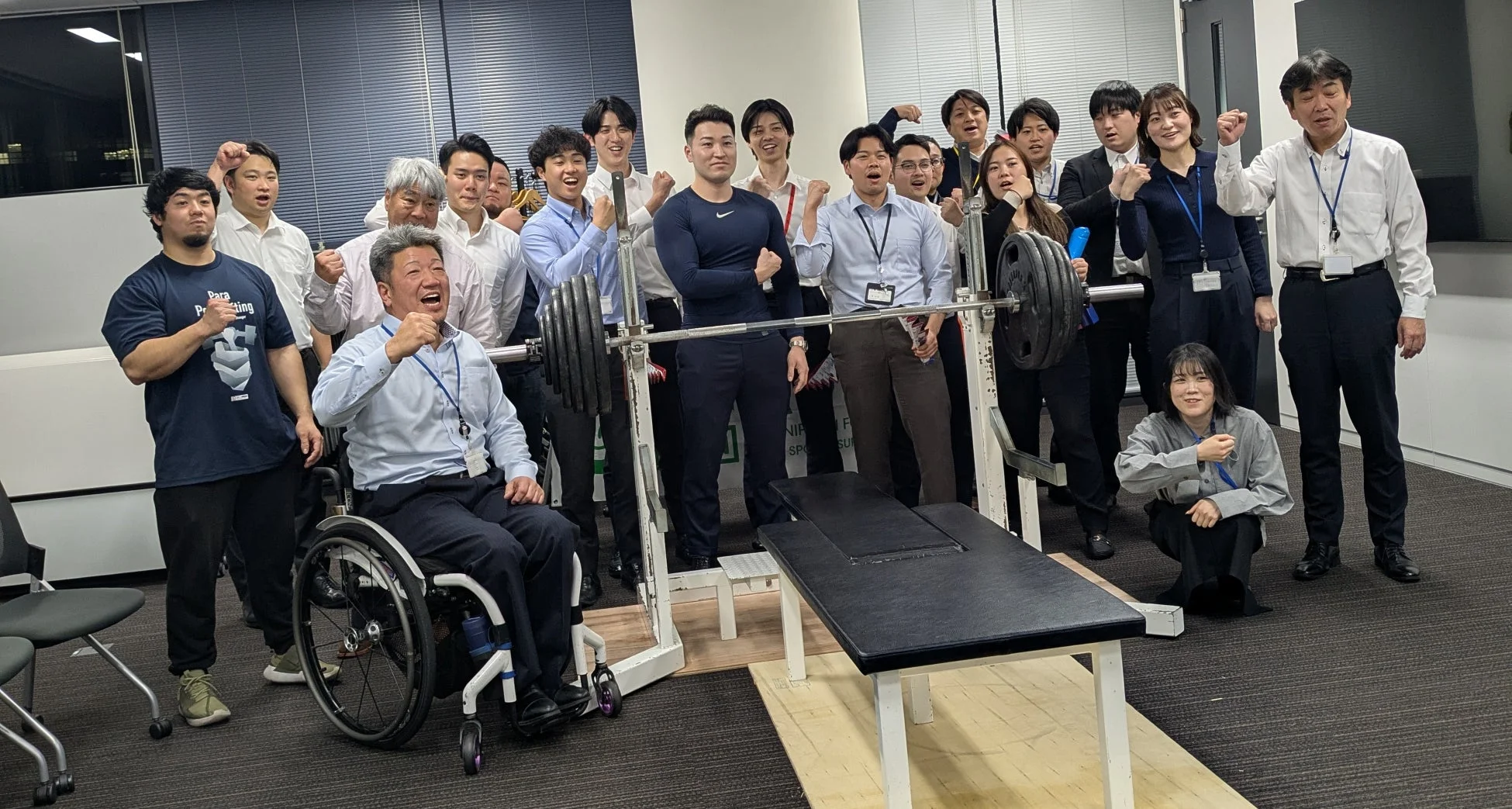 オフィス内でベンチプレスを囲んで応援する社員たち