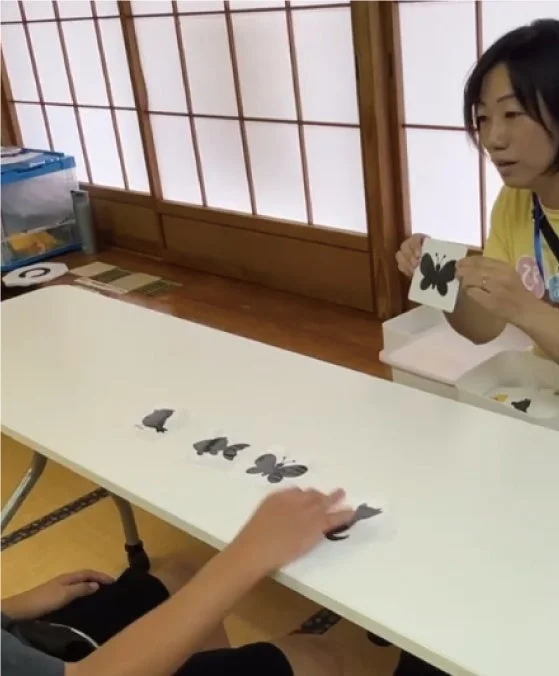 女性が卓上で蝶のシルエットカードを使った活動を行っている。向かい側の人物がテーブル上のカードに手を伸ばしており、学習や発達支援のような様子が見られる。背景は障子の和室。
