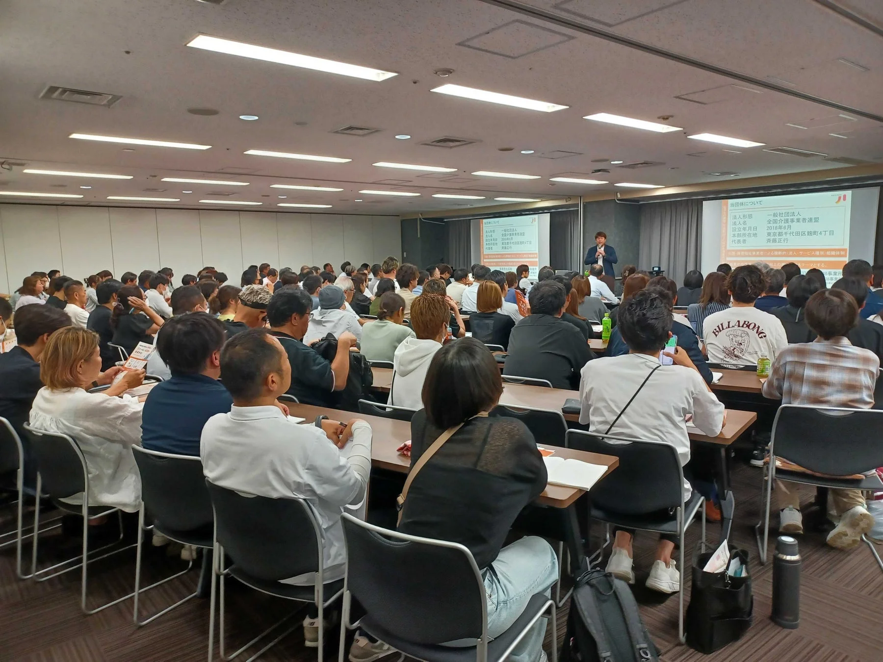 多くの参加者が集まる会場で、一人の講師がスクリーンを使ってプレゼンテーションを行っています。