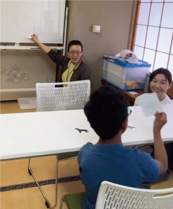 和室のような空間で、3人の人々がテーブルを囲んで学習やワークショップを行っている様子です。一人の女性がホワイトボードの前で説明し、別の女性は笑顔で座っています。テーブルには犬のシルエットの絵があり、参加者の一人がカードを持っています。
