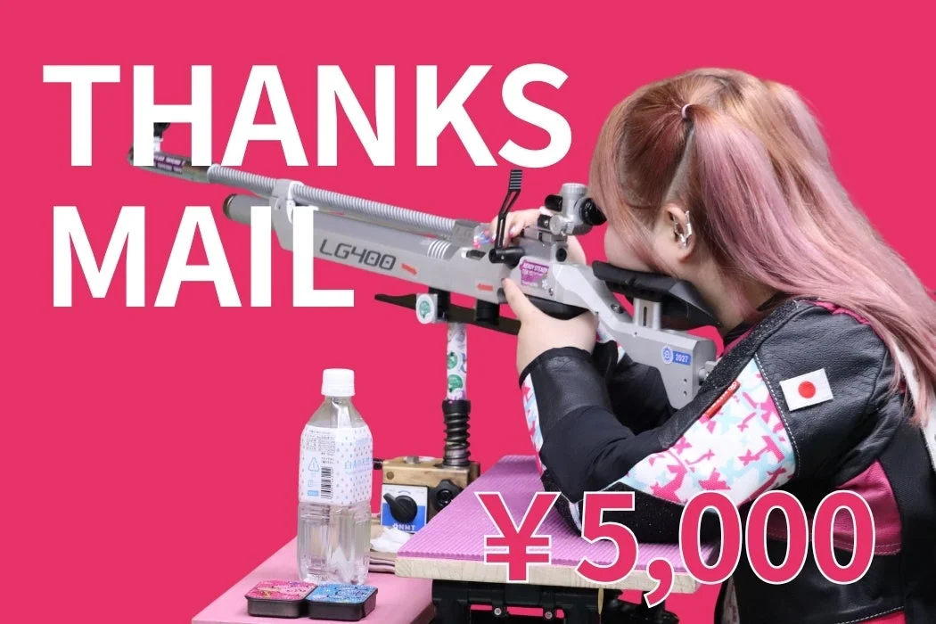 ピンク色の背景で、ピンク髪の女性が日本国旗のパッチ付き射撃ジャケットを着てエアライフルを構えている。画像には「THANKS MAIL」と「¥5,000」の文字がオーバーレイされている。