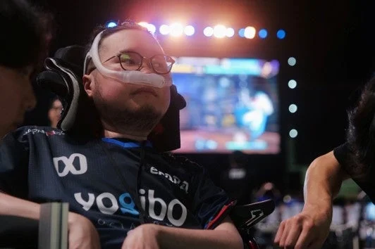 eスポーツイベントのような会場で、鼻カニューレを装着し眼鏡をかけた男性が、競技用のユニフォームを着て車椅子に座っている。集中した表情で前を見つめている。