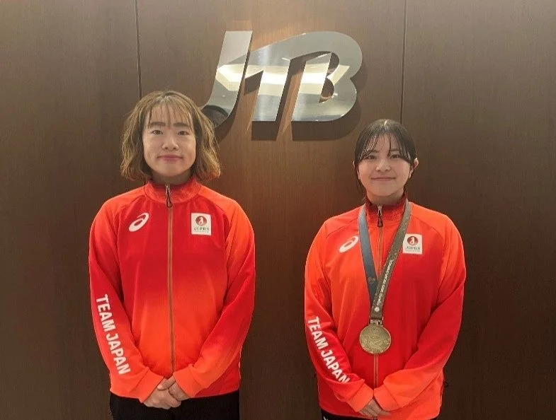 JTBのロゴを背景に、赤とオレンジのグラデーションの「TEAM JAPAN」のユニフォームを着た二人の女性が立っています。一人の女性は金メダルを首にかけており、二人とも笑顔で写っています。